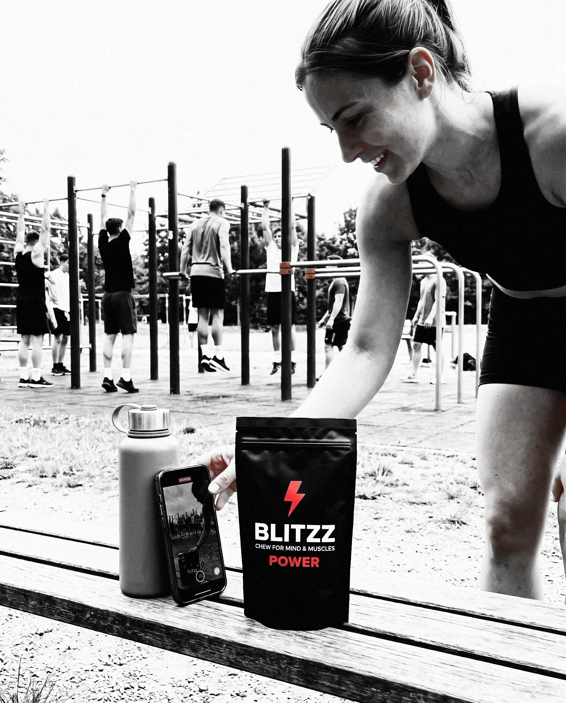 An athlete in an outdoor calisthenics park checking the training clip she filmed on her phone, a pack of BLITZZ POWER on the bench